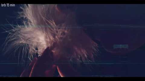 Video of a person with long, messy hair and a blurred face, standing in a dimly lit room with horizontal lines of light. The image has a gritty, moody atmosphere.