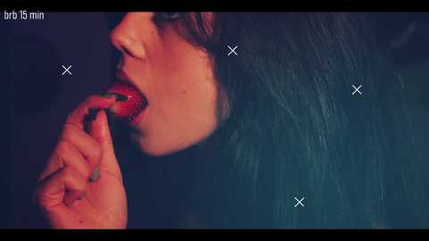 Video of a woman with long, dark hair, pale skin, and full lips, licking a red strawberry. The background is blurred and dark, with white x marks overlaying the image.