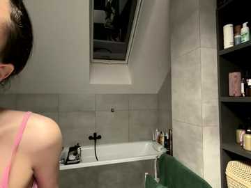 Video of a woman in a pink tank top facing away from the camera in a modern bathroom with beige tiles, a green bathtub, and a black shelf holding toiletries.