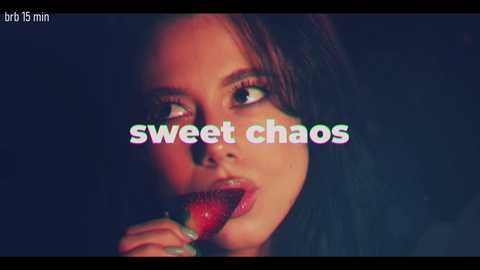 Video of a woman with fair skin and dark hair, holding a red strawberry near her lips. Text overlay reads \"sweet chaos\" in pastel colors. Dark background.