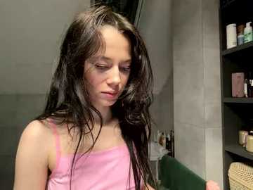 A young girl with long, wet black hair, wearing a pink tank top, stands in a bathroom with gray walls, a green towel, and a shelf filled with toiletries.