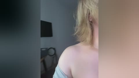 Video of a blonde woman with fair skin, wearing a light grey off-shoulder top, partially visible in a dimly lit room with a black TV and a black chair in the background.
