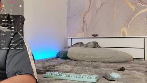 A video shows a person in a gray shirt lying on a plush, gray bed with a light blue keyboard and mouse nearby. The background features a white wall with abstract art.