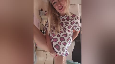 A video of a smiling young woman with blonde hair, wearing a leopard print top, lifting her leg, in a cluttered, pink-walled room.