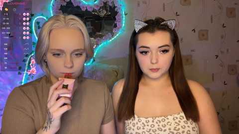 Video of a blonde woman with a tattoo, wearing a beige top, eating a strawberry, and a brunette woman with a leopard-print top, both in a room with a neon heart and floral wallpaper.