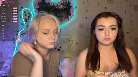 Video of two young women with fair skin, one with short blonde hair, the other with long brown hair. They're sitting indoors, surrounded by a pink heart-shaped neon light and floral wallpaper.