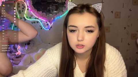Video of a young woman with fair skin and long brown hair, wearing a white knitted sweater, and glittery cat ears. Background features colorful neon lights and scattered papers.
