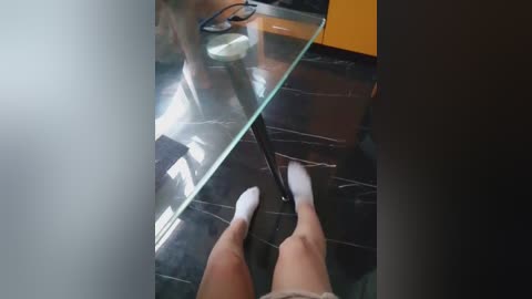 Video of a person's feet in white socks, standing on a polished black marble floor, holding a glass door frame, with a blurry reflection of a person in the background.
