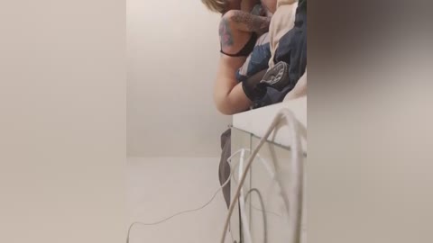 A video of a woman with short blonde hair, wearing a dark blue dress, leaning against a white wall. She has tattoos on her left arm. A tangled white cord is visible on the floor.