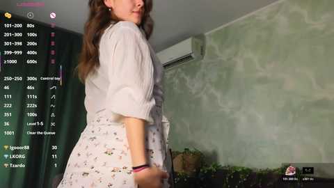 A video of a woman with wavy brown hair, wearing a white floral dress, standing in a dimly lit room with green walls. The image includes text overlays from a social media app.