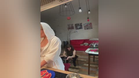 A video captures a woman in a white robe standing at a kitchen counter, while another woman in a black dress sits on a sofa in a modern living room with red cushions and hanging lights.