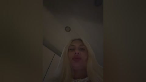 A dimly lit video of a blonde woman with fair skin, wearing a white veil, standing in a narrow, dark corridor. The image is grainy, suggesting a low-quality camera or poor lighting.