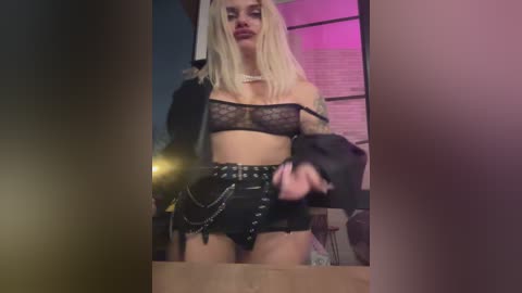 Video of a blonde woman with tattoos in a black lace bra and high-waisted shorts, holding a jacket, in a dimly lit room with purple lighting.