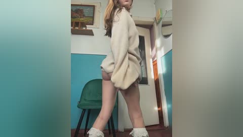 Video of a young woman with light skin, long blonde hair, and a white, oversized sweater, standing in a teal-walled room with a green chair and framed painting in the background.