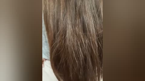 Close-up video of a woman's long, straight, brown hair, with strands slightly parted in the center, partially covering a light blue shirt. Background is blurred, focusing attention on the hair texture and color.