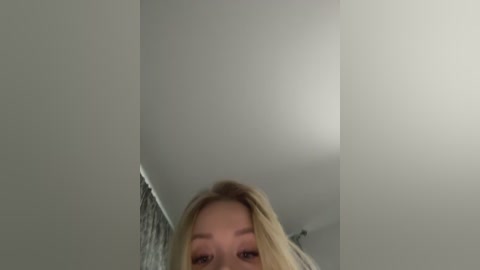 Video of a young woman with long blonde hair, partially visible from the neck up, against a plain white ceiling. She has fair skin and light makeup.