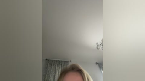 A video of a blonde woman in a light-colored room with beige walls and a silver chandelier. The background shows a patterned curtain and part of a white ceiling.