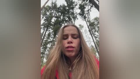 A video captures a young woman with long, blonde hair, wearing a red top, standing in a forest with tall trees, her eyes closed, and a serene expression.