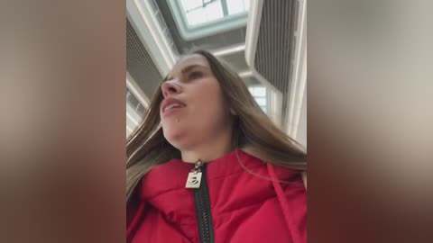 Video of a young woman with light skin and long brown hair, wearing a red jacket with a black zipper, standing in an indoor space with large windows and metallic vent covers.