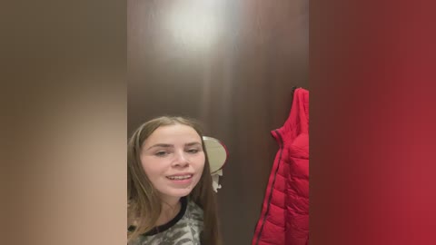 Video of a young Caucasian woman with straight, light brown hair, smiling, wearing camouflage-patterned clothing. She stands in a small, dimly lit room with a red jacket hanging on the right wall.