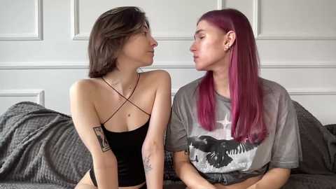 Video of two women with tattoos, one with black hair and a black halter top, the other with purple hair and a grey graphic T-shirt, sitting on a gray couch.