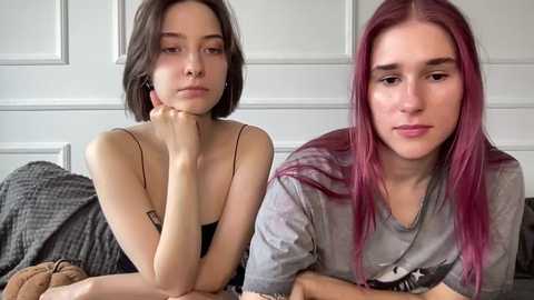 Video of two young women with short hair, one with pale skin and dark hair, the other with fair skin and pink hair, sitting on a bed, looking contemplative.