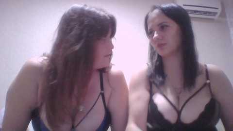 Video of two Caucasian women with long, straight dark hair, wearing black lace lingerie with strappy details, standing close together in a dimly lit room with white walls and an air conditioner.