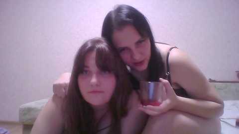 Video of two young women with long dark hair, one topless, the other in a black top, sitting on a bed in a dimly lit room with light-colored walls.
