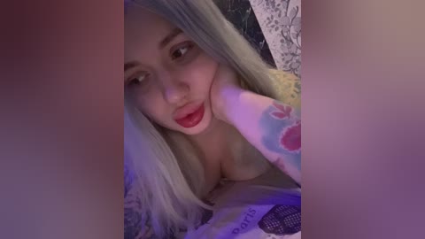 Video of a pale-skinned woman with long, platinum blonde hair and red lipstick, lying on a bed, partially covered by a soft, white blanket. Her face is close to a pink, heart-shaped object, casting a soft glow.