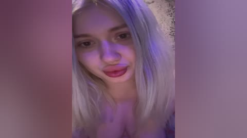 Video of a young woman with long, platinum blonde hair, wearing red lipstick and a black top. Her face is partially obscured by purple lighting.