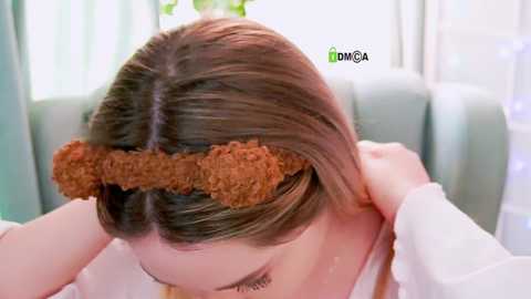 Video of a woman with straight, light brown hair, wearing a curly brown hair extension headband. She's dressed in a white top, indoors with light-colored curtains in the background.