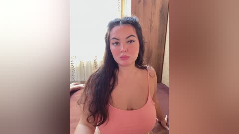 Video of a young woman with long dark hair, wearing a pink spaghetti-strap top, sitting on a wooden floor in a sunlit room with beige walls and a radiator.