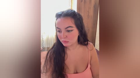 Video of a young Latina woman with long, wavy black hair, fair skin, and a slender physique, wearing a pink spaghetti-strap top. She's indoors near a window with sheer curtains, in a cozy, wooden room.