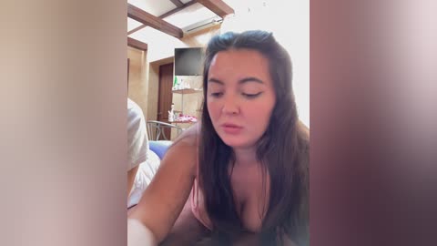 A video of a young woman with long brown hair, wearing a pink top, leaning forward. The background shows a bed, wooden ceiling, and a TV.