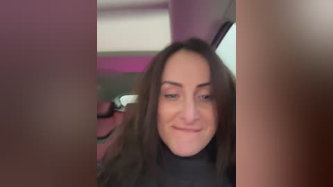 A video of a young woman with long brown hair and light skin, smiling slightly, seated in a car with pink and white interior.