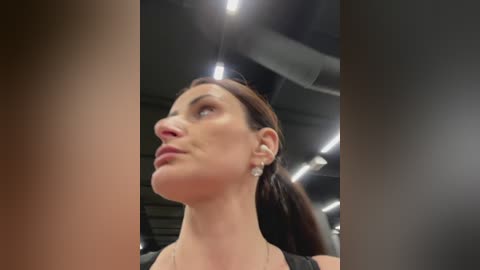 Video of a woman with fair skin, brown hair tied back, wearing a black top and pearl earrings, looking upward in an industrial setting with fluorescent lights.