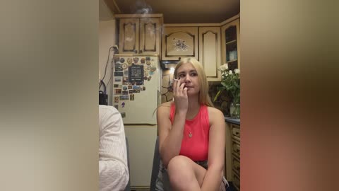 Video of a blonde woman in a red tank top, seated in a kitchen with floral wallpaper, vintage cabinets, and a cluttered refrigerator.