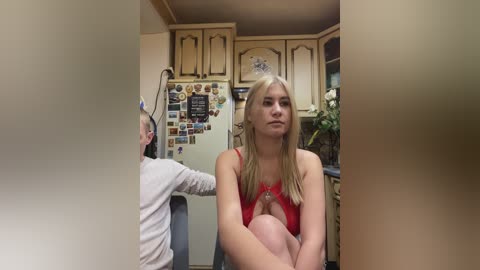 Video of a blonde woman with fair skin, wearing a red spaghetti-strap top, sitting in a kitchen. Another person is behind her, partially obscured.