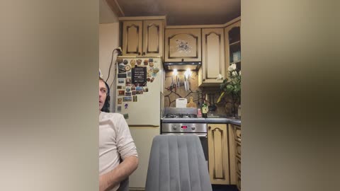 Video of a man with a black beard and white shirt, standing in a vintage kitchen with beige cabinets, a fridge covered in magnets, and a gray chair.