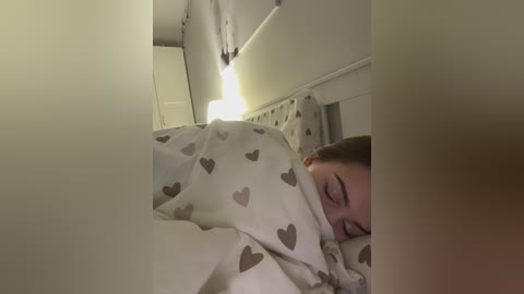 A video of a woman with light skin, brown hair, and closed eyes, sleeping under a white blanket with heart patterns, in a dimly lit room with a white ceiling and a light fixture.