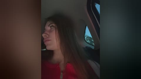 A video of a young woman with long brown hair, wearing a red top, gazing out a car window at night, with dimly lit, blurred trees outside.