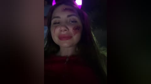 Video of a young woman with pale skin, long brown hair, wearing a red top, and purple devil horns, with smeared makeup including red around her eyes and mouth. Dimly lit background suggests an indoor Halloween party.