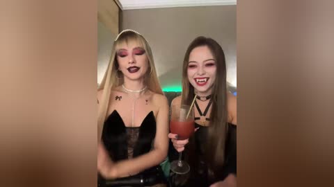 Video of two women with long blonde and brunette hair, wearing dark, form-fitting dresses, holding red drinks, smiling at the camera.
