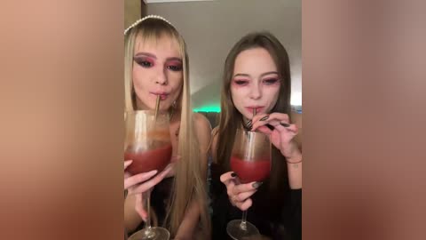Video of two young women with long hair, wearing black tops, sipping red drinks through straws, with heavy makeup and a blurred background.