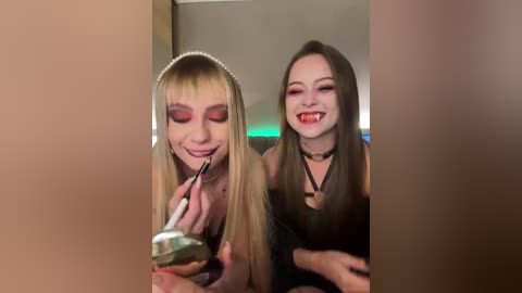 Video of two young women with long brown hair, one wearing a headband and dramatic red eye makeup, the other with a black choker and red lipstick, applying makeup.