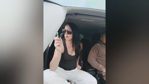 Video of a woman with long black hair, wearing sunglasses, a black tank top, and white pants, sitting in the back seat of a car, holding a phone, with another person partially visible.