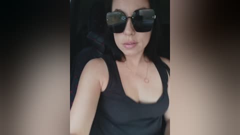 A video of a woman with medium skin tone, wearing large black sunglasses and a black tank top, sitting in a car. She has a neutral expression.