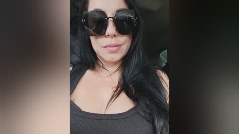 Video of a woman with long black hair, wearing black sunglasses and a black tank top, standing indoors, blurred background.