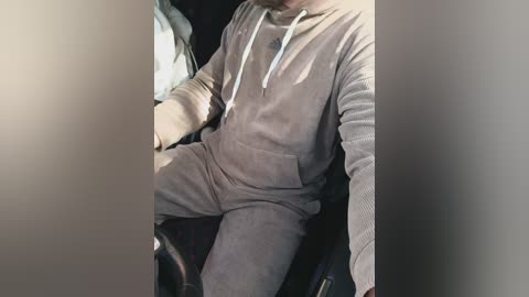 Video of a person in a gray, relaxed-fit hoodie and matching pants, sitting in a car, hands on the steering wheel.