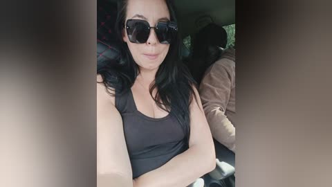 Video of a woman with long black hair, wearing large dark sunglasses, a black tank top, and sitting in a car.